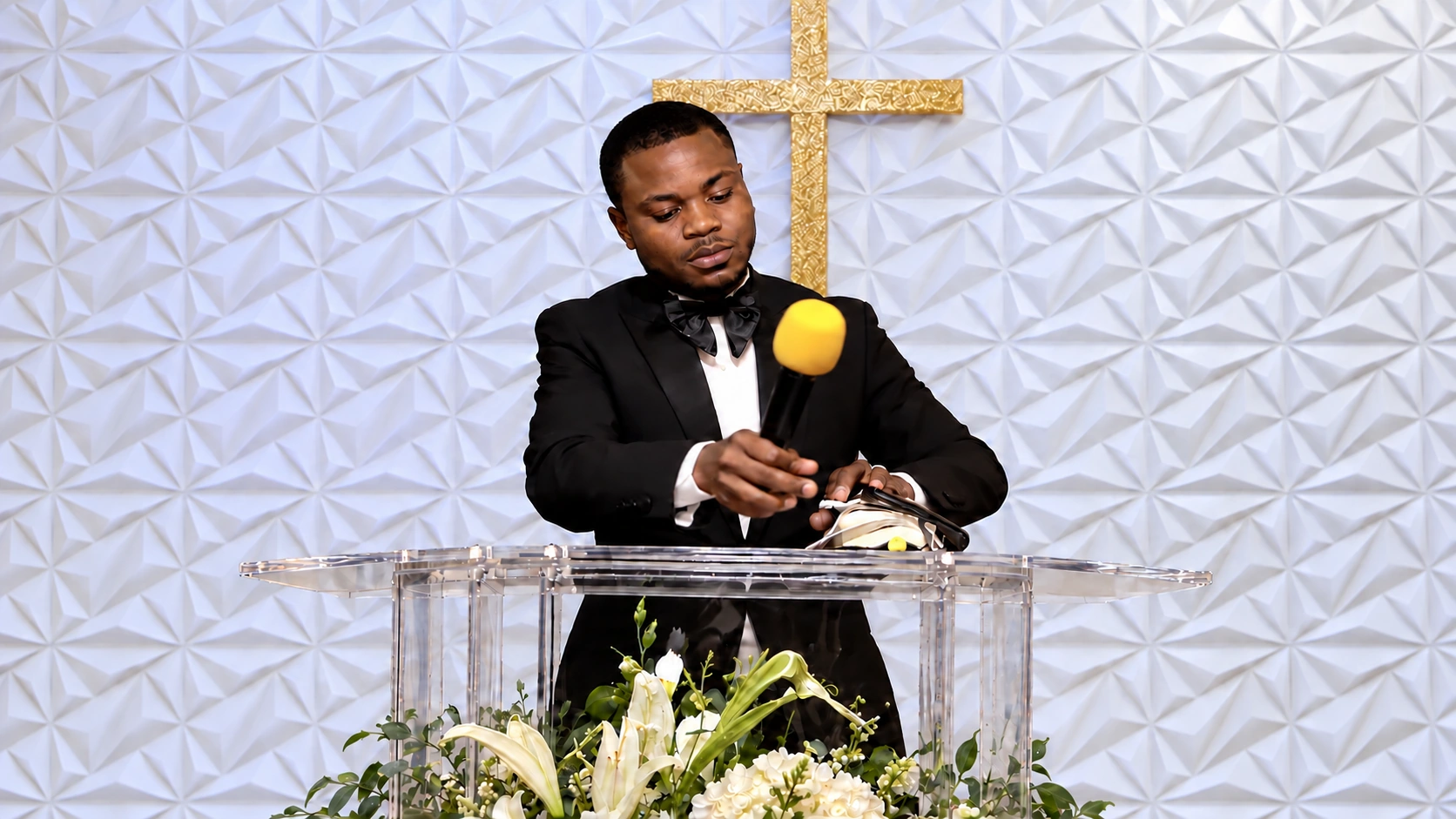 A young speaker in a black bow tie holding a yellow microphone at the transparent pulpit. — Artistically restyled surroundings